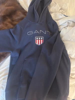 Mörkblå hoodie från GANT - Mörkblå hoodie från GANT med broderad logotyp och ett snyggt emblem med stjärnor och ränder på bröstet. Hoodien har en stor huva och en klassisk magficka. Materialet är mjukt och bekvämt, perfekt för chill dagar. Made in Turkey.