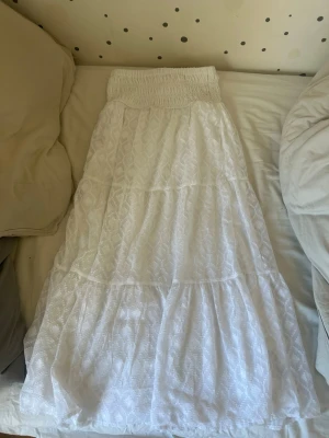 Vit långkjol med smock och mönster - Säljer en vit långkjol med bred smock i midjan och diskret geometriskt mönster. Kjolen har flera lager och är luftig med lätt struktur i tyget, perfekt för varma dagar. Materialet känns som bomull och kjolen har en feminin och avslappnad look.