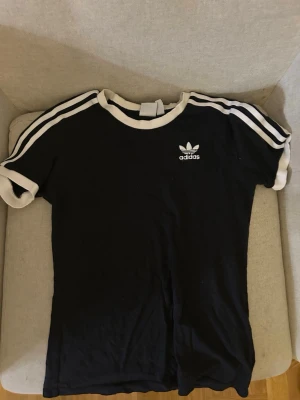 Svart Adidas t-shirt med vita detaljer - Cool svart t-shirt från Adidas med vita ribbade kanter och klassiska vita stripes på axlarna. Loggan är tryckt på bröstet och materialet är mjuk bomull. Perfekt för dig som gillar sportig stil och vill ha något bekvämt och snyggt.