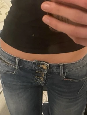 Blå low waist jeans från MOD - Säljer ett par blå low waist jeans från MOD med tre sneda knappar i gylfen och en unik dragkedjedetalj vid fickan. Jeansen har slitningar och kontrastsömmar som ger en cool och trendig vibe. Perfekta för dig som gillar detaljer och streetstyle.