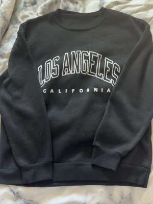 Svart sweatshirt Los Angeles tryck - Svart sweatshirt med stort vitt tryck där det står 'Los Angeles California' på bröstet. Tröjan har rund halsringning, ribbade muddar vid ärmslut och nederkant. Perfekt för en avslappnad och trendig streetwear-look