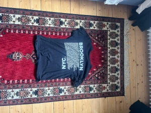 Svart t-shirt med Brooklyn-tryck - Cool svart t-shirt med stort vitt Brooklyn-tryck på ryggen och NYC-text. Klassisk rund hals och korta ärmar. Perfekt för dig som gillar streetstyle och urban känsla. Materialet är mjuk bomull som är skönt att bära hela dagen.