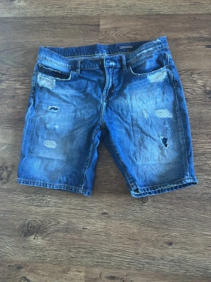 Blå jeansshorts Tommy Hilfiger - Säljer ett par blå jeansshorts från Tommy Hilfiger med slitna och trasiga detaljer. Klassisk femficksdesign, normal passform och tillverkade i bomull. Perfekt för dig som vill ha en cool och avslappnad stil under sommaren.