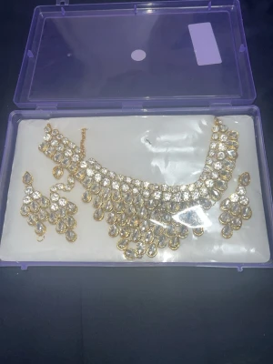 Guldigt halsband med pärlor och kristaller - Säljer ett elegant halsband i guldfärg med droppformade pärlor och gnistrande kristaller. Halsbandet har en lyxig design med flera lager och matchande örhängen. Perfekt för dig som vill sticka ut med ett statement-smycke. Levereras i en lila plastask.