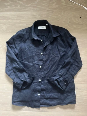 Svart linneskjorta från Les Deux - Säljer en svart skjorta från Les Deux i linne med klassisk krage och vita knappar. Skjortan har långärmad design och en stilren look som passar perfekt till jeans eller chinos. Materialet är lätt och luftigt, vilket gör den skön att bära.