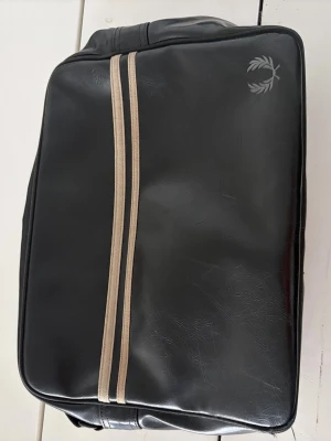 Svart axelväska från Fred Perry - Cool svart axelväska från Fred Perry med två beige ränder på framsidan och klassisk logotyp i hörnet. Väskan har en rektangulär form och justerbar axelrem. Materialet är konstläder som ger en stilren och sportig look.