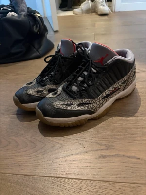 Air Jordan 11 Low svart/grå sneakers - Säljer ett par Air Jordan 11 Low sneakers med svart läder och grå elefantmönster på ovandelen. Skorna har röda detaljer på tungan och insidan, samt vita och beige sulor. Klassisk design med snörning och låg profil. OBS! PRIS KAN DISKUTERAS