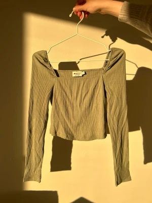 Ribbad offshoulder topp från NA-KD - Trendig ribbad offshoulder topp från NA-KD i en ljus olivgrön nyans. Toppen är croppad med långa ärmar och har en smickrande, figurnära passform. Perfekt att styla med höga jeans eller kjol!