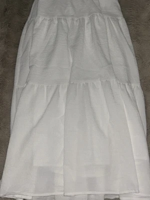 Vit långkjol med volangdetaljer - Säljer en vit långkjol i ett strukturerat tyg med bred resår i midjan och volangdetaljer. Kjolen har en luftig och flowy känsla, perfekt för varma dagar. Materialet känns som bomull eller frotté och kjolen har en enkel, stilren look.