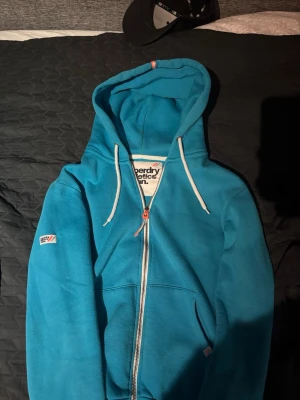 Blå zip hoodie från Superdry - Snygg blå hoodie från Superdry med dragkedja framtill och vita snören i huvan. Märkeslogga på ärmen och tryckt etikett i nacken. Mjuk bomullskänsla och två fickor fram. Perfekt för en avslappnad streetwear-look.