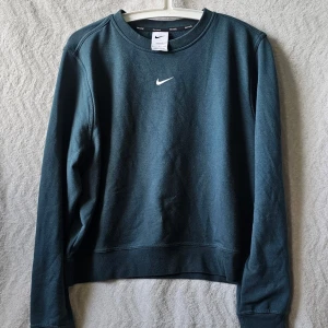 Mörkgrön Nike sweatshirt Dri-FIT - Snygg mörkgrön sweatshirt från Nike med Dri-FIT material som håller dig torr och bekväm. Tröjan har rund halsringning, långa ärmar och en vit Nike-logga på bröstet. Perfekt för dig som gillar sportig och stilren design.