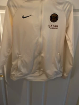 Beige PSG Nike ziptröja med huva - Snygg beige ziptröja från Nike med PSG-logga och Qatar Airways-tryck på bröstet. Tröjan har huva, dragkedja framtill och två fickor. Perfekt för träning eller när du vill representera ditt favoritlag.