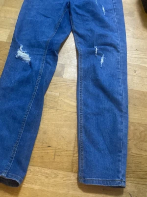 Blå slitna mom jeans - Säljer ett par blå mom jeans med flera slitningar på benen för en trendig och avslappnad look. Jeansen har hög midja och raka ben. Perfekta för dig som gillar en casual och cool stil. Materialet är klassisk denim.