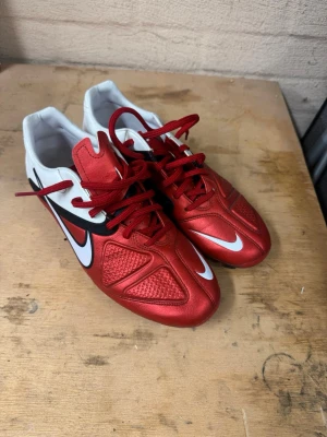 Nike röda och vita fotbollsskor - Snygga fotbollsskor från Nike i rött och vitt med svarta detaljer och klassisk Swoosh-logga på sidan. Skorna har snörning och en modern design med förstärkta partier framtill för bättre bollkontroll. Perfekta för dig som vill sticka ut på planen.