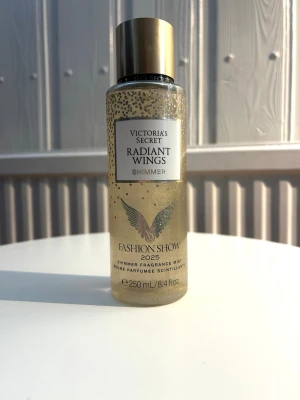 Victoria's Secret Radiant Wings Shimmer - En lyxig shimmer mist från Victoria's Secret, Radiant Wings Shimmer, Fashion Show 2025 limited edition. Flaskan är genomskinlig med guldfärgade detaljer och lock, fylld med skimrande guldpartiklar. Doften har inslag av glitter magnolia, krämig amber och drömmiga toner. Bara testad på huden, säljer eftersom jag inte tycker att doften passade mig🩷
