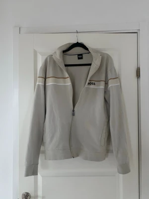 Beige hoodie med dragkedja från BOSS - Snygg beige hoodie från BOSS med dragkedja och hög krage. Den har vita och bruna detaljer över bröstet samt BOSS-loggan på vänster sida. Hoodien är långärmad och har ribbade muddar. Perfekt för en chill och stilren look.