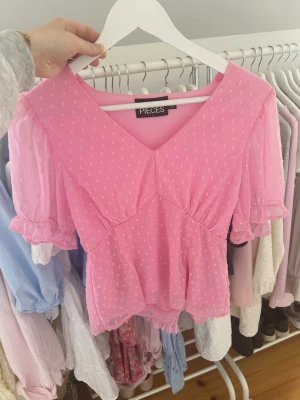 Rosa blus med v-ringning från Pieces - Söt rosa blus från Pieces med v-ringning och korta puffärmar. Blusen har ett diskret prickigt mönster och volangdetaljer vid ärmslutet. Perfekt för dig som gillar en feminin och trendig stil.