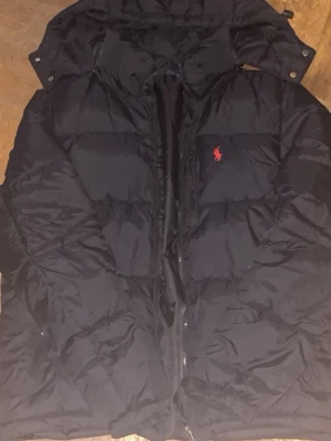 Ralph lauren jacka svart - Säljer en svart pufferjacka från Ralph Lauren med röd broderad logga på bröstet. Jackan har huva, dragkedja framtill och är vadderad för extra värme. Perfekt för kalla dagar och har en klassisk, sportig look.