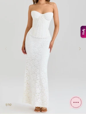 Vit spetsklänning med corsettopp - Elegant långklänning i vit spets med axelbandslös corsettopp och figurnära passform. Klänningen har vacker spetsdetalj över hela och markerad midja som framhäver siluetten. Perfekt för speciella tillfällen och sommarens fester.