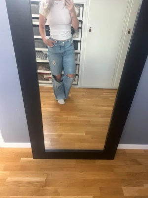 Blå slitna wide leg jeans - Säljer ett par blå wide leg jeans med stora slitningar på knäna och en mindre reva på låret. Jeansen har hög midja och klassisk femficksdesign. Perfekt för dig som gillar en avslappnad och trendig look.