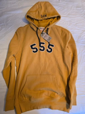 Gul hoodie med 555-tryck - Snygg gul hoodie från Triple 5 Soul med svart-vita siffror '555' broderade på bröstet och svart dragsko i huvan. Klassisk känguruficka framtill och ribbade muddar vid ärmar och nederkant. Perfekt för en avslappnad streetwear-look.
