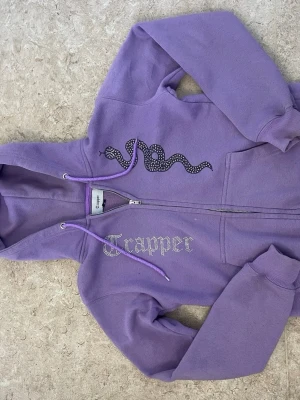 Lila hoodie med orm och text - Lila hoodie från Trapper med dragkedja, huva och fickor framtill. På bröstet finns en svart orm i strasstenar och texten 'Trapper' i glittriga stenar. Mjuk bomullsmix och ribbade muddar vid ärmar och nederkant. Perfekt för en chill och trendig look.