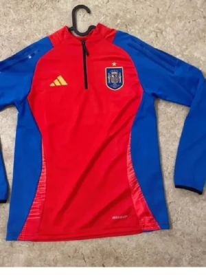 Röd och blå träningsjacka Adidas Spanien - Säljer en röd och blå långärmad träningsjacka från Adidas med Spaniens landslagsmärke på bröstet. Jackan har halv dragkedja framtill, gul Adidas-logga och detaljer i blått längs ärmar och sidor. Materialet är syntetiskt och passar perfekt för sportaktiviteter.