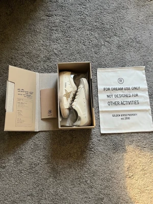 Golden Goose vita sneakers med glitterstjärna - Säljer min syrras Golden Goose sneakers i vitt läder med beige detaljer och en glittrig guldstjärna på sidan. Skorna har en låg profil, beige snörning och silverfärgad häl. Snygg design med klassisk siluett och coola detaljer. Allt på bilderna kommer med,även Golden goose passport. Passar 37/36