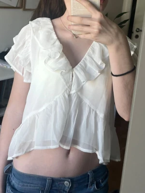 Vit volangblus från Nelly - Säljer en vit blus från Nelly med v-ringning och volangdetaljer. Blusen är gjord i ett tunt, transparent material och har korta ärmar. Jättefin till sommaren!! Den är aldrig använd