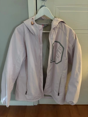 Ljusrosa vindjacka från OTP - Ljusrosa vindjacka från OTP med huva och dragkedja framtill. Jackan har två sidofickor med dragkedja och ett stort tryck på bröstet. Materialet är lätt och passar perfekt för blåsiga dagar. Snygg och sportig design med praktiska detaljer. Storlek 46 men ganska liten i storlek.