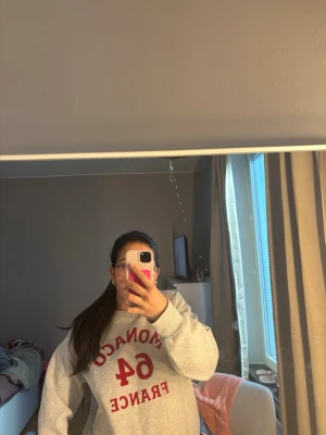 Grå sweatshirt med röd text - Cool grå sweatshirt med röd text 'MONACO 64 FRANCE' på bröstet. Tröjan har en klassisk rund hals och långa ärmar, perfekt för en avslappnad och trendig look. Materialet ser ut att vara mjukt och bekvämt, idealiskt för chill dagar.