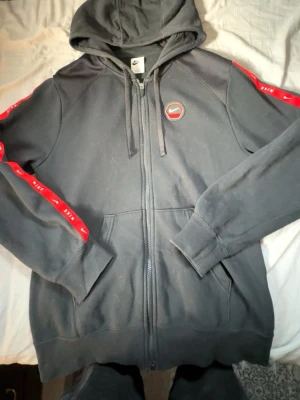 Svart Nike hoodie med dragkedja - Svart hoodie från Nike med dragkedja framtill och huva med snören. Röda detaljer med Nike-logga längs ärmarna och ett Nike-märke på bröstet. Stor Nike-tryck på ryggen. Fickor framtill. Knappt använd, blivit för stor. Priset kan diskuteras!