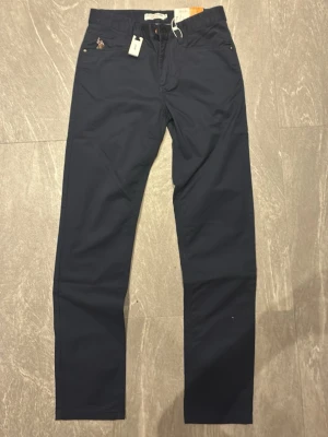 Mörkblå slim chinos från U.S. Polo Assn. - Snygga mörkblå chinos från U.S. Polo Assn. i slim fit-modell. Byxorna har klassisk design med raka ben, bälteshällor och diskret logga vid fickan. Tillverkade i ett mjukt och bekvämt material som passar perfekt till en modern stil.