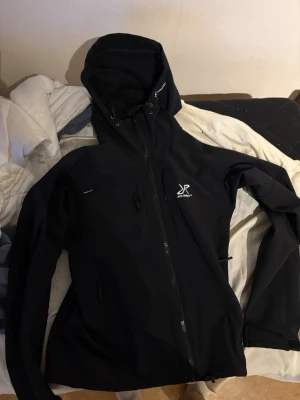 Svart vindjacka RevolutionRace  - Svart vindjacka från RevolutionRace med huva och dragkedja framtill. Jackan har flera fickor med dragkedja och justerbar huva. Tillverkad i slitstarkt material som passar perfekt för utomhusaktiviteter. Stilsäker logga på bröstet.