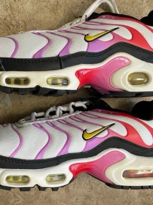 Nike Air Max Plus TN rosa/vit - Nike Air Max Plus TN sneakers med vit mesh, rosa och röda detaljer, svart kant och gula Swoosh-loggor. Synliga Air-bubblor i sulan och cool vågdesign på ovandelen. Perfekt för dig som vill sticka ut med färg och stil.