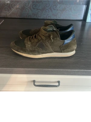 Olivgröna sneakers med mockadetaljer - Säljer ett par olivgröna sneakers med snygga mockadetaljer och meshpartier. Skorna har klassisk låg profil, snörning och en diskret logga på sidan. Passar perfekt till jeans och har en stilren look med jordnära färger.