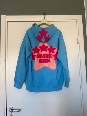 Blå hoodie från Aelfric Eden - Säljer en blå hoodie från Aelfric Eden med stor rosa stjärna och text på både fram- och baksidan. Baksidan har dessutom flera rosa stjärnor och huvan är dekorerad med stjärnor. 