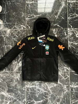 Svart Brasilien Nike vindjacka - Cool svart vindjacka från Nike med Brasilien landslagslogga på bröstet och flera sponsortryck i gult och orange. Jackan har huva, dragkedja och fickor. Perfekt för dig som gillar sport och streetstyle.