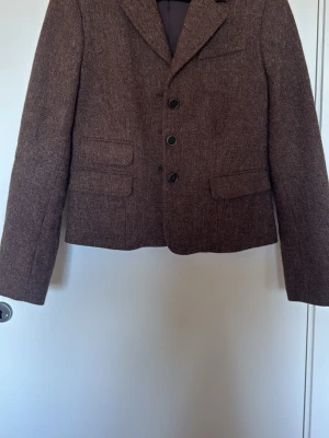 Brun kavaj med tre knappar - En klassisk brun ullkavaj med tre svarta knappar framtill och två fickor med lock. Kavajen har rak passform, lång ärm och stilren look. Perfekt för dig som gillar tidlösa plagg och vill ha något som funkar till många outfits. Kvalitativt märke, sparsamt använd med fint foder.