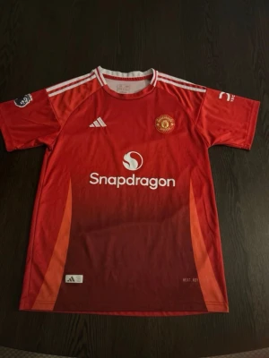 Manchester United röd matchtröja Adidas - Röd Manchester United matchtröja från Adidas med vita detaljer och klubbens emblem på bröstet. Tröjan har Snapdragon-logga på framsidan och är tillverkad i lätt, ventilerande material. Perfekt för fotboll och träning.