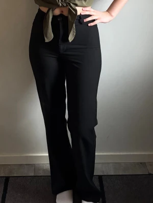 Svarta kostymbyxor med raka ben - Snygga svarta kostymbyxor med hög midja och raka ben. Byxorna har klassisk knappstängning och är tillverkade i ett slätt material som ger en stilren look. Perfekta för dig som vill ha en tidlös och trendig byxa.