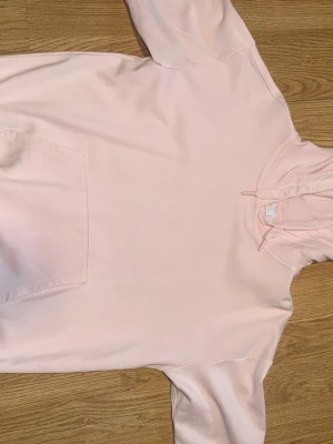 Ljusrosa hoodie med huva - En enkel och stilren ljusrosa hoodie med huva och känguruficka framtill. Hoodien har dragsko i halsen och ribbade muddar vid ärmslut och nederkant.