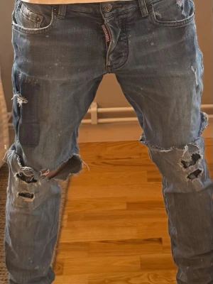 DSQUARED2 jeans  - Säljer ett par blå DSQUARED2 jeans i stl 14Y med kraftigt slitna detaljer och stora hål på knäna. Jeansen har en klassisk femficksdesign och är gjorda i denim. De har en rak passform och råa kanter vid hålen för en cool och avslappnad look.