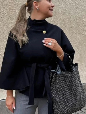 Missäy poncho - Stilren svart cape-kappa med hög krage och dekorativ guldfärgad knapp. Kappan har vida ärmar och knytband i midjan som ger en snygg siluett. Perfekt för att skapa en elegant look. Materialet ser ut att vara mjukt och följsamt.