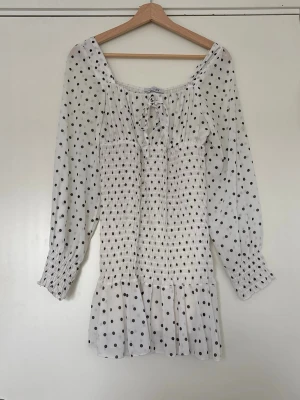 Vit prickig kortklänning från Design by Si - Söt vit kortklänning från Design by Si med svarta prickar, smockad midja och volang nertill. Klänningen har långa ärmar och en knytning vid halsen. 