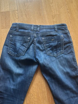 Blå Miss Sixty jeans med kontrastsömmar - Snygga blå jeans från Miss Sixty Collection med kontrastsömmar och klassiska femficksdetaljer. Modellen har en cool knapp med mönster och tydlig logotyp på bakfickan. Perfekt för dig som gillar trendiga och bekväma jeans.
