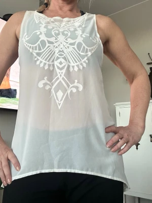 Vit ärmlös blus med mönster - Säljer ett vitt genomskinligt linne med snyggt broderat mönster framtill. Linnet har rund halsringning. 