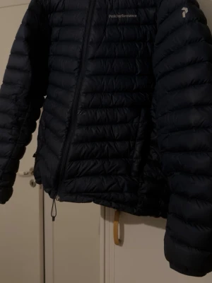Mörkblå dunjacka från Peak Performance - Säljer en svart dunjacka från Peak Performance med quiltad design och dragkedja framtill. Jackan har lång ärm och logotyp på bröstet samt ärmen. Perfekt för kalla dagar och har en stilren look som passar till många outfits.