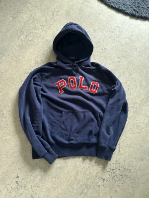 Marinblå hoodie från Polo Ralph Lauren - Snygg marinblå hoodie från Polo Ralph Lauren med stor röd POLO-text i frotté på bröstet. Klassisk känguruficka framtill, ribbade muddar och justerbar huva. Perfekt för en avslappnad och trendig look.