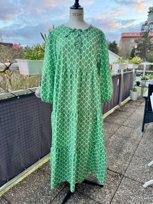 Grön långklänning med mönster - Långklänning i grönt och vitt. Klänningen har lång ärm, rund halsringning med knyt och en avslappnad passform. Perfekt för vår och sommar, tillverkad i ett lätt och luftigt material.